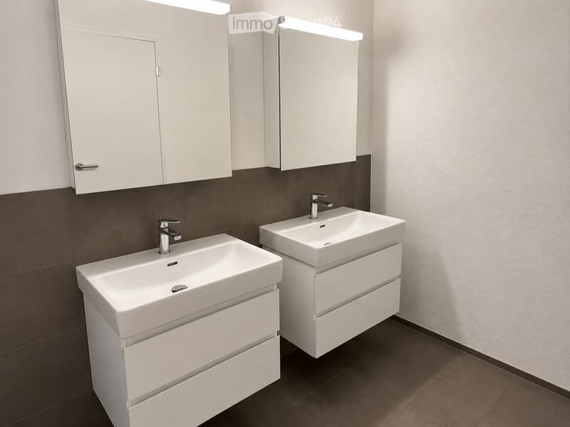 Modern bathroom with two white sinks, each with a chrome faucet, integrated cabinets, and illuminated mirrors above.