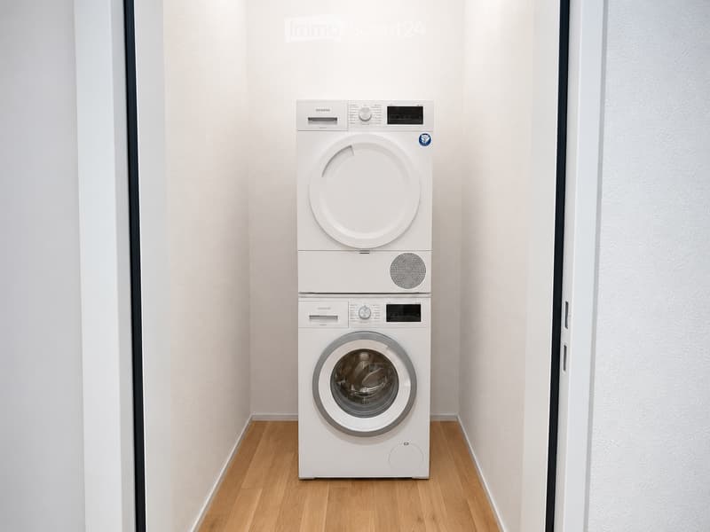 A small, well-lit room with light-colored walls and wooden flooring, featuring a stacked washing machine and dryer unit.