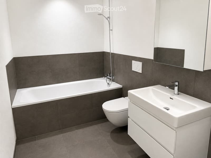 Modern bathroom with a white bathtub, toilet, sink, and mirror. Dark grey tiles on walls and floor, sleek fixtures.