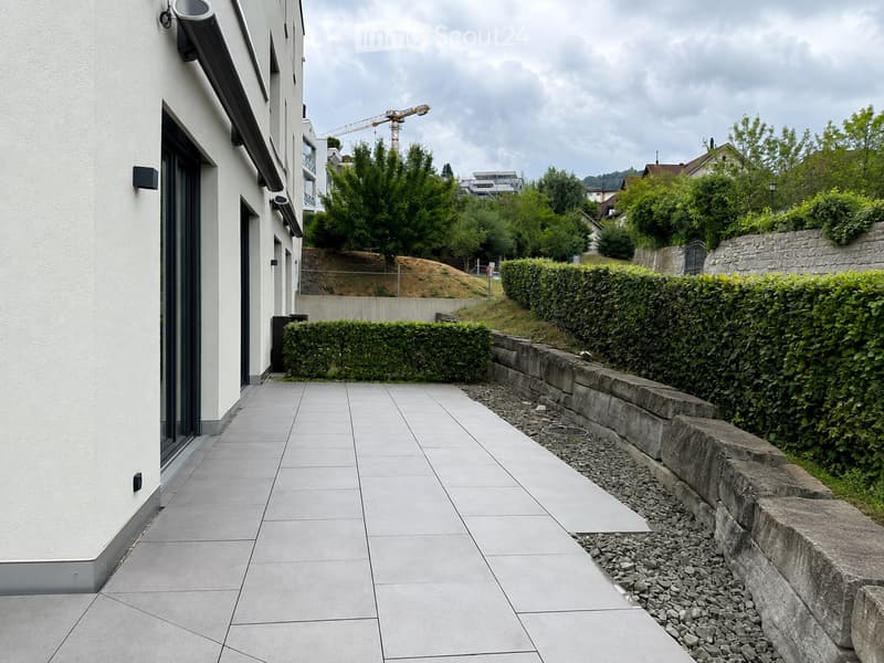 A paved outdoor area adjacent to a modern building, featuring a stone retaining wall, neatly trimmed hedges, and a gravel bed.
