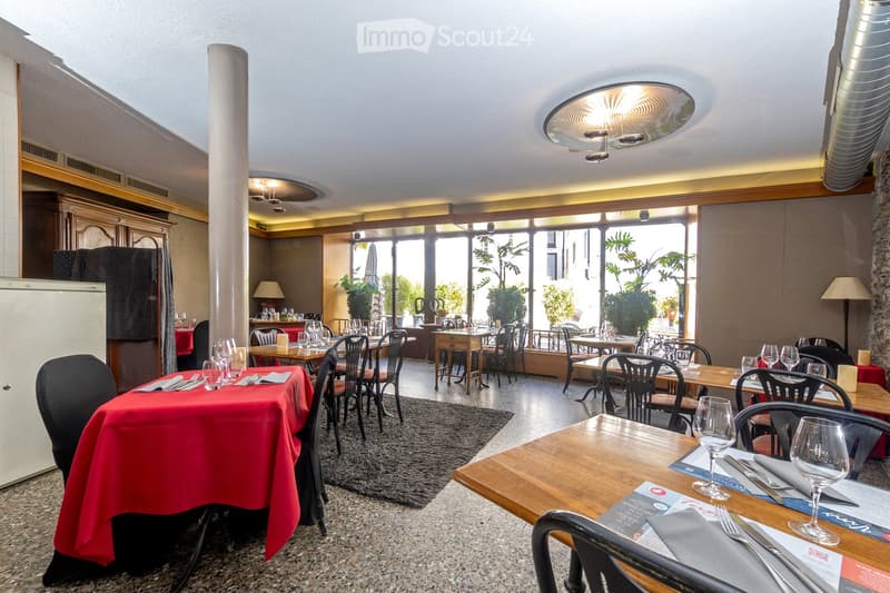 Restaurant interior with multiple tables and chairs, wine glasses, cutlery, and napkins set on tables, large windows with outdoor view, and a refrigerator in the background.