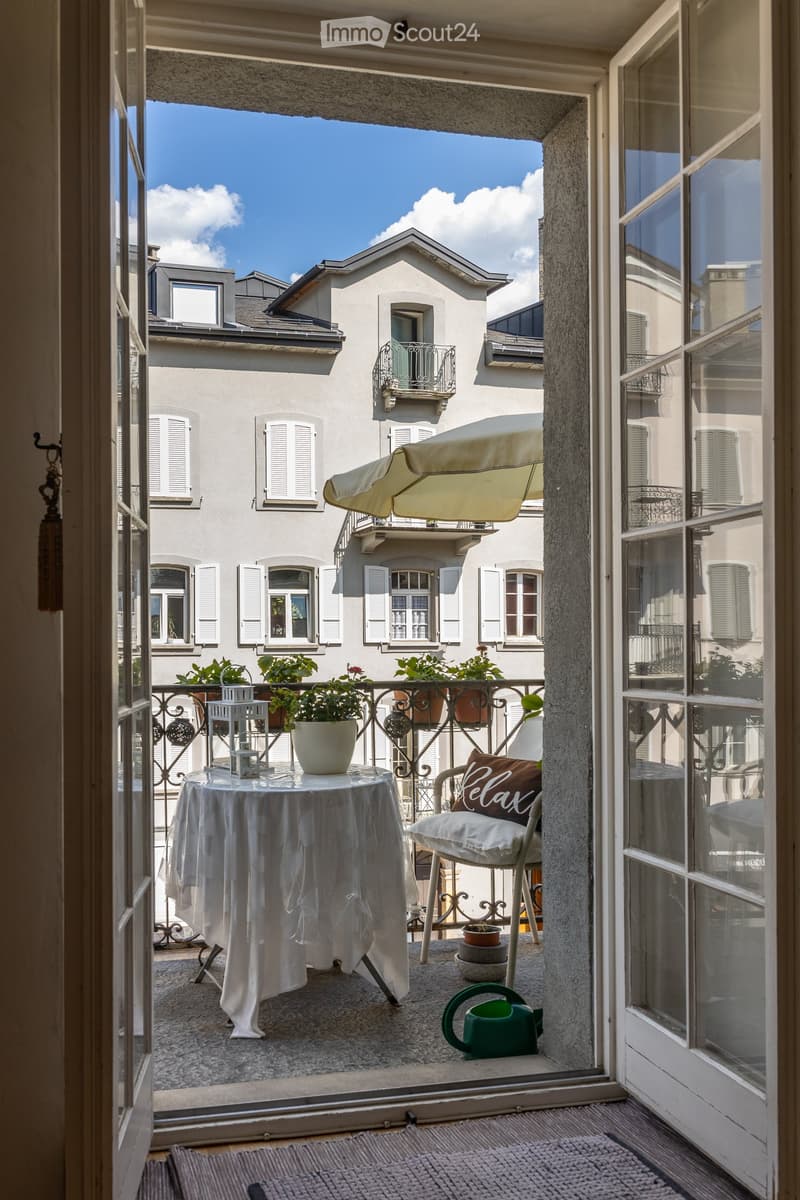 Balcony with a table, chair, potted plants, umbrella, and watering can, overlooking a cityscape with traditional buildings.