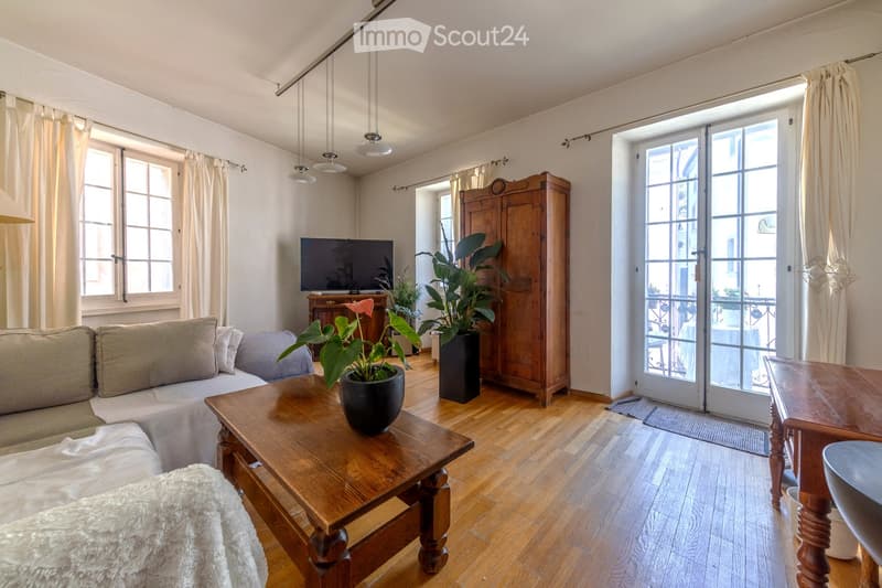 Bright living room with wooden flooring, large windows, and sliding glass doors leading to a balcony. Features a sofa, coffee table, television, armchair, and potted plants.