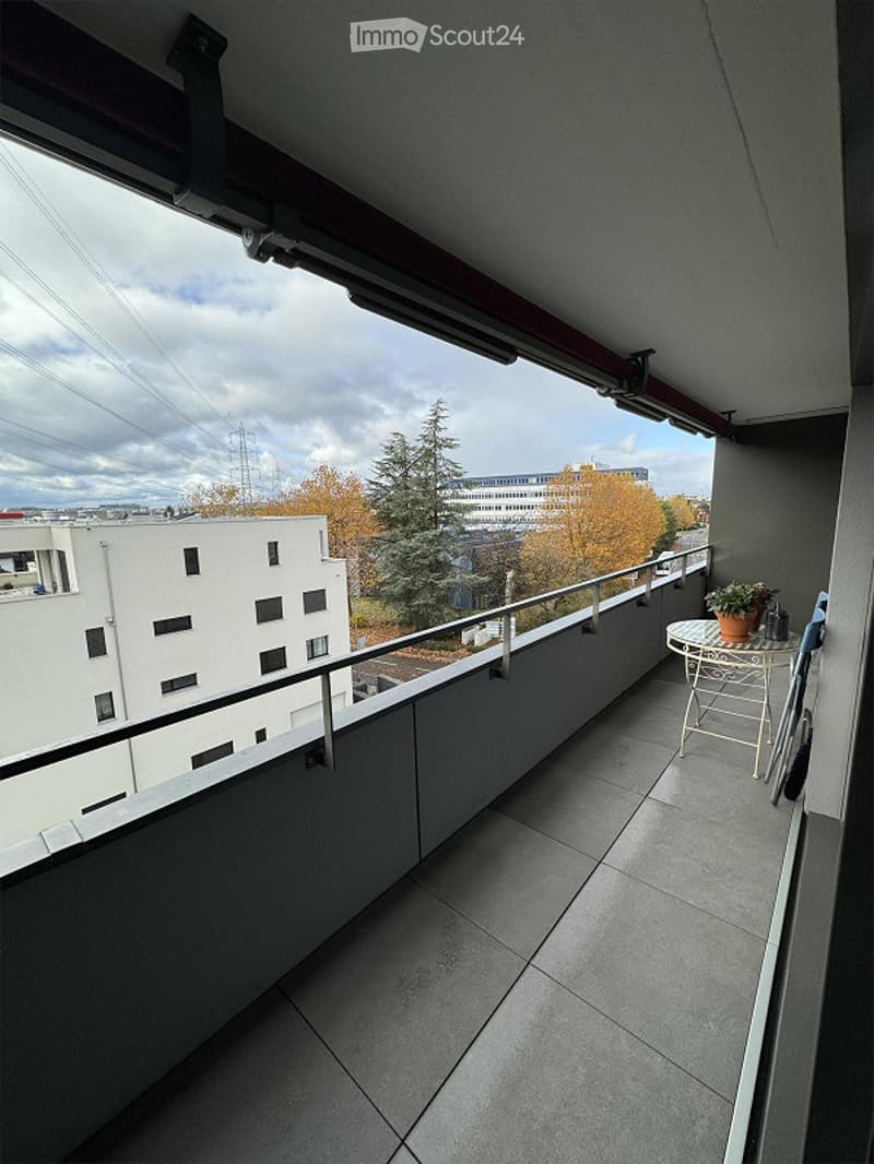 Balcony with tiled floor, metal railing, small round table, chair, and flower pot. Offers a view of surrounding buildings and trees.