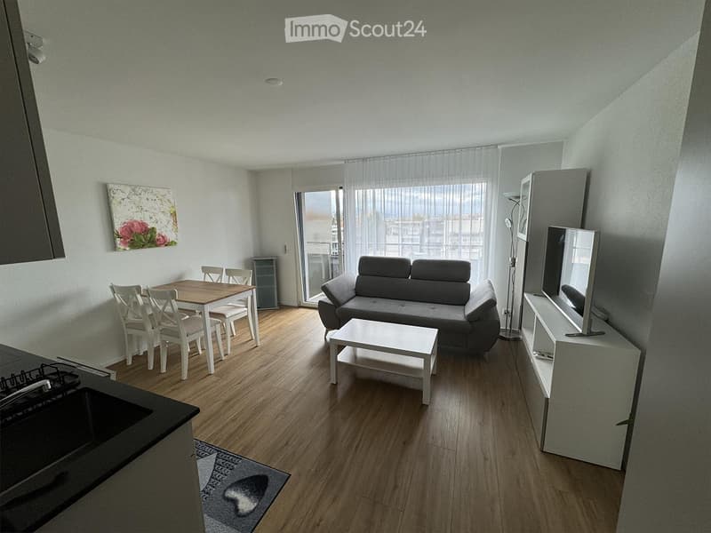 Modern open-plan living and dining area with wooden flooring, grey sofa, white coffee table, dining table with chairs, TV stand, and a view through large windows.