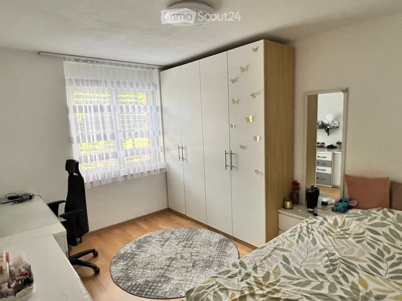 A tidy bedroom with a bed, a chair, a large wardrobe, a mirror, a round table, and a small stool. The room has wooden flooring and a window with sheer curtains.