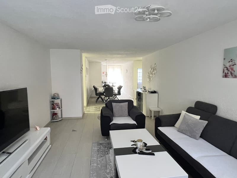 Modern living room with a grey sofa, coffee table, dining area with chairs and table, TV screen, and decorative shelf.