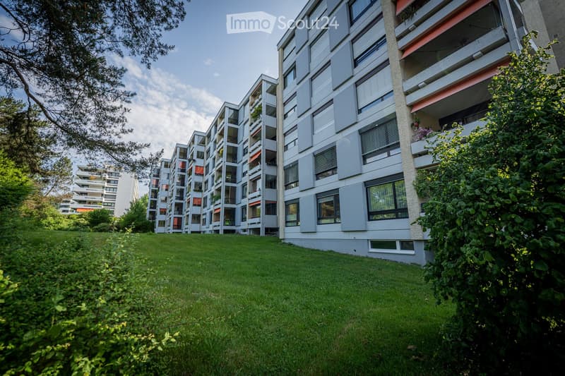 Modern multi-story apartment building with balconies and large windows, surrounded by greenery and a well-maintained lawn.