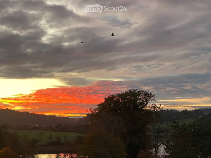 Scenic view of a sunset over a valley with fields, trees, and a body of water reflecting the sky.