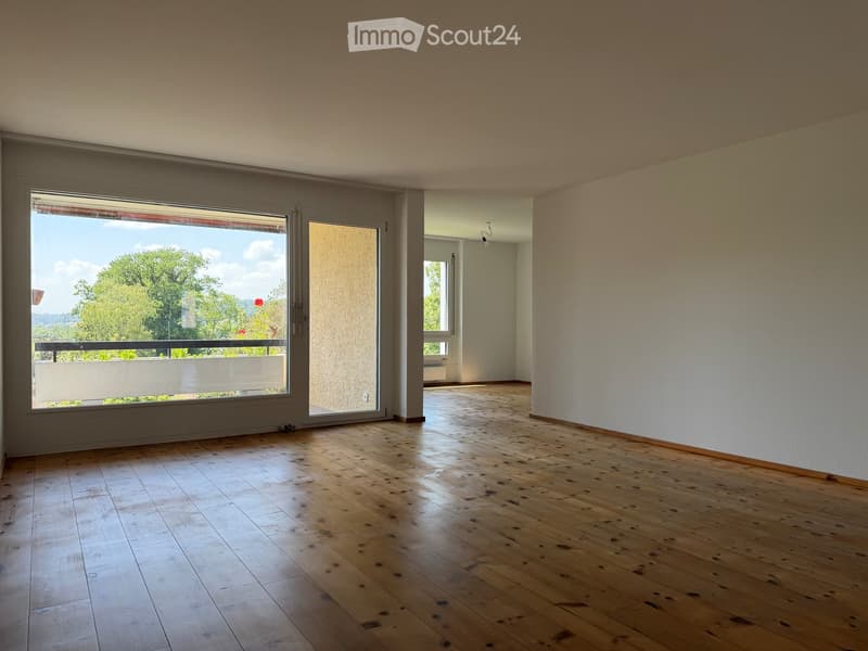 Empty room with wooden flooring, large windows, and a sliding glass door leading to a balcony with a view of greenery.