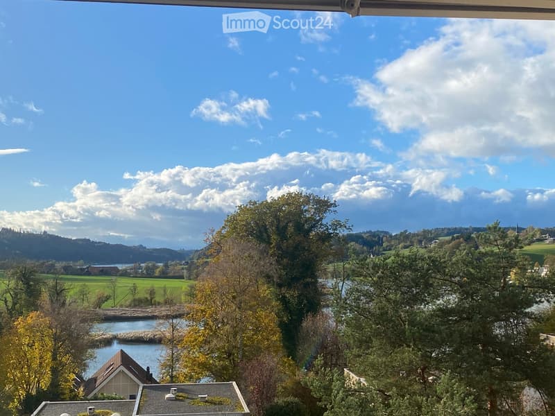 View from a balcony or terrace overlooking a scenic landscape with trees, a body of water, and distant hills under a partly cloudy sky.