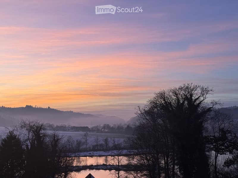 A serene landscape view at sunrise or sunset, featuring a calm river reflecting the sky, surrounded by bare trees and distant hills.