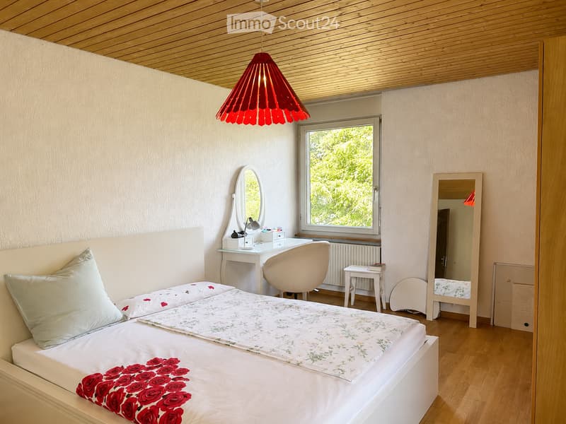 Cozy bedroom with a white bed, floral bedding, a wooden ceiling, a large window with a view of greenery, a desk with a chair, and a mirror.