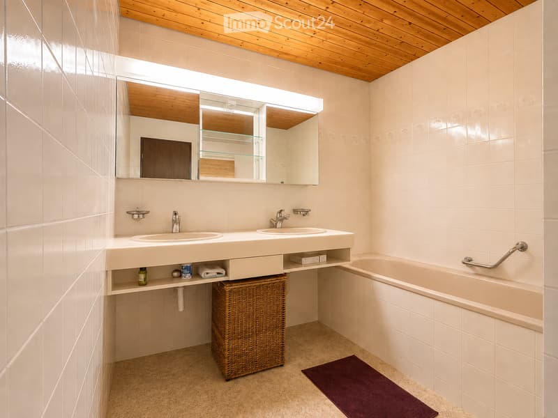 Modern bathroom with white tiled walls, a double sink vanity, a large mirror, a bathtub with a grab bar, and a wicker basket under the sink.