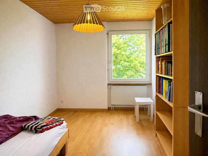 Minimalist bedroom with wooden flooring, a single bed with colorful bedding, a small white table, a wooden bookshelf filled with books, and a modern pendant light fixture.
