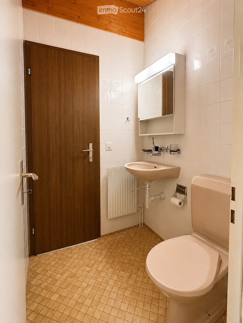 Modern bathroom with white tiled walls, beige tiled floor, a toilet, a sink with a mirror cabinet, and a radiator. The door is wooden with a silver handle.