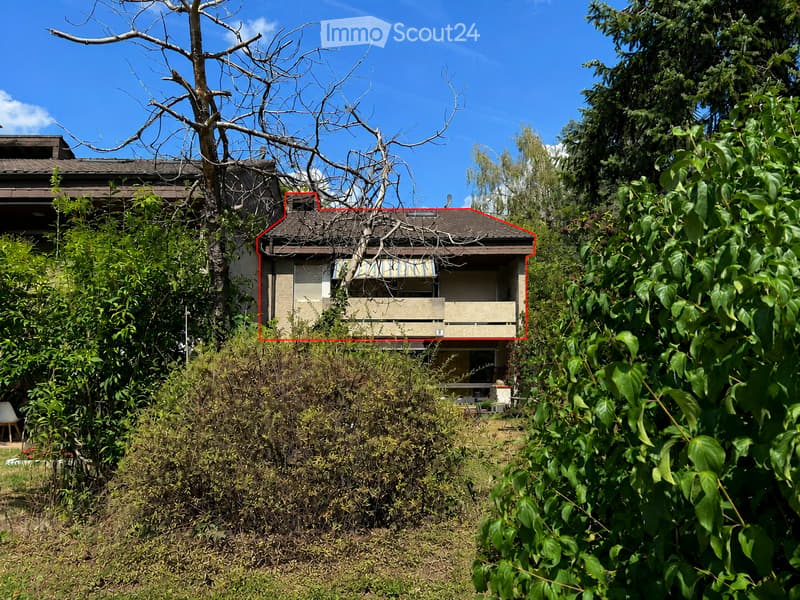 A two-story house with a visible balcony and a covered patio area, surrounded by overgrown vegetation and trees.