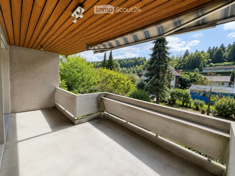 Balcony with wooden ceiling, striped awning, concrete floor, and railing, overlooking a green, hilly landscape with houses and trees.