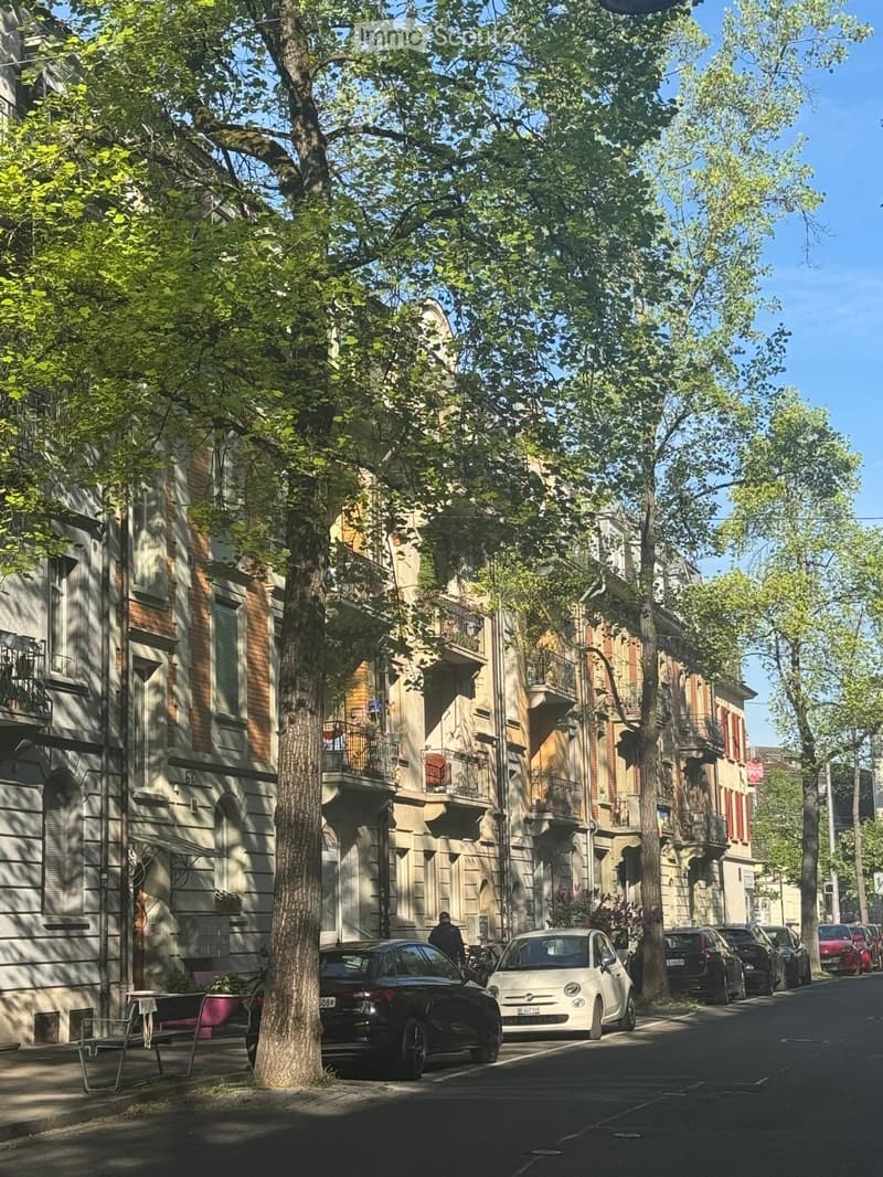 A tree-lined street with multi-story residential buildings, balconies, parked cars, and a few pedestrians.