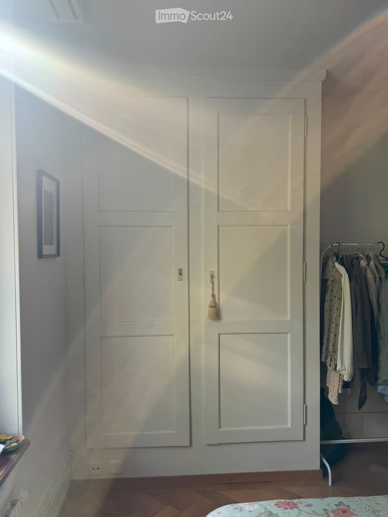 A well-lit hallway with two closed doors, a small table with a mirror, and a wardrobe with hanging clothes.