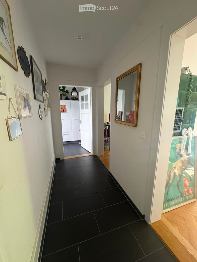 Hallway with black tiled floor, white walls, framed pictures, a large mirror, and an open door leading to another room with white cabinets and a clock on top.