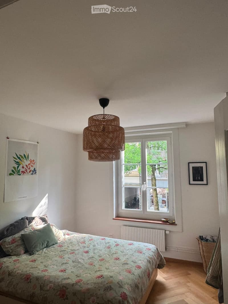 Cozy bedroom with a wooden floor, a large window with a view, a bed with a floral duvet, a pendant light, and a picture frame on the wall.