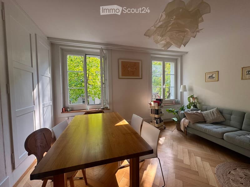 Bright living room with wooden flooring, large windows, a sofa, a dining table with chairs, a floor lamp, and decorative items.