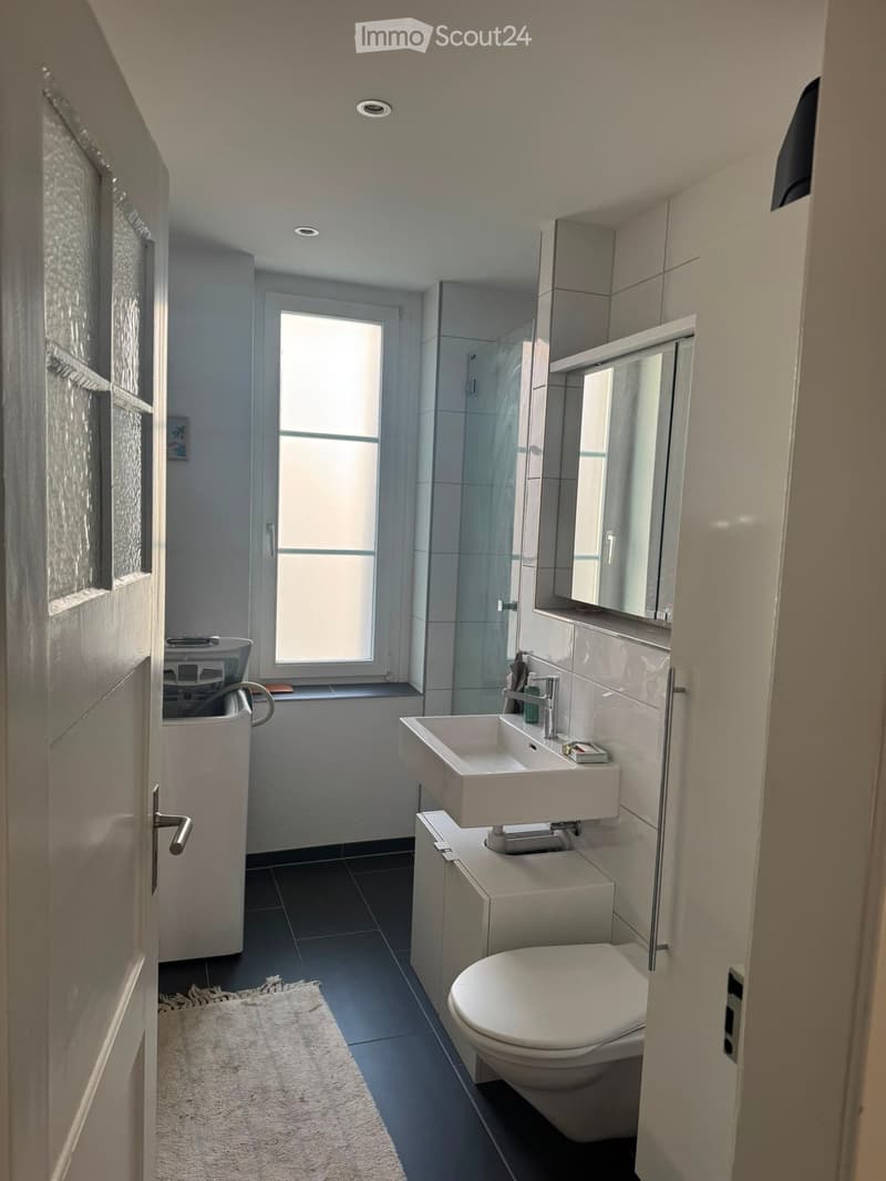 Modern bathroom with white sink, toilet, mirror, and shower. Features dark tiled floor, white cabinetry, and a large window providing natural light.