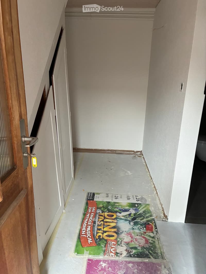 A narrow hallway with white walls, a wooden door on the left, and a partially visible poster on the floor.
