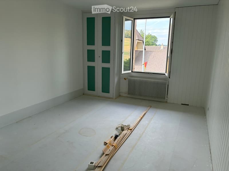 Empty room with white walls, wooden floor, large window with view, radiator, and some construction materials on the floor.