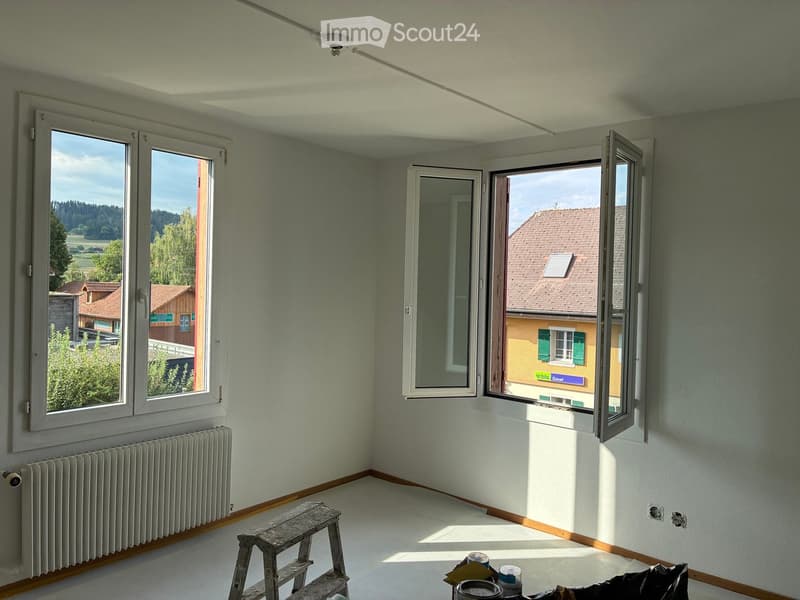 Empty room with light grey walls, wooden baseboards, white ceiling, and large windows offering a view of the countryside and neighboring houses. A radiator is visible under one window, and a small wooden stool and paint cans are placed on the floor.