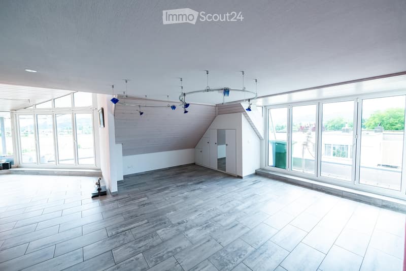 Bright, empty room with wooden flooring, large windows, and modern lighting fixtures. The space features white walls and a slanted ceiling typical of an attic or loft area.