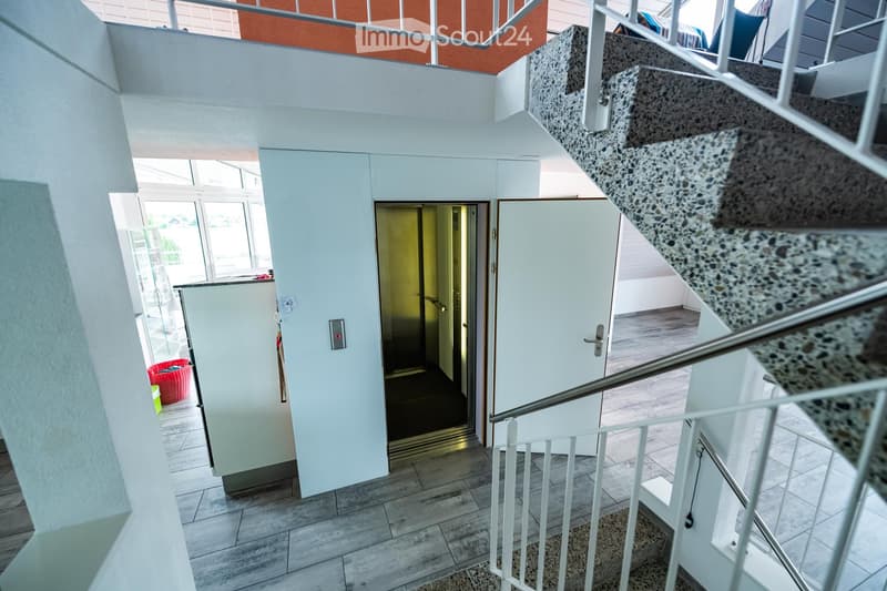 Modern hallway with a staircase leading upwards, white walls, tiled floor, and a partially visible red basket in the background.