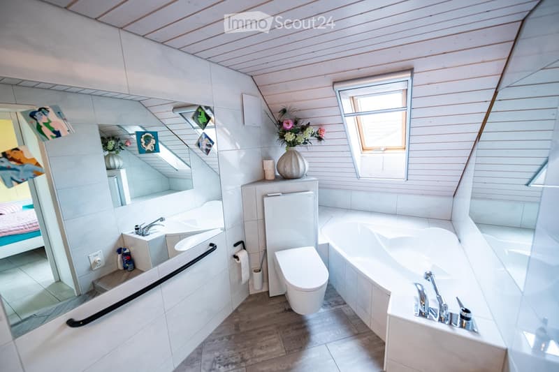 Modern bathroom with a large bathtub, toilet, sink, and mirror. The room features white tiled walls, wooden flooring, and a skylight window providing natural light. A vase with flowers and a few decorative items are placed on the windowsill.