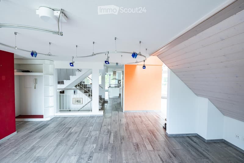 Open-plan living space with wooden flooring, white walls, and modern lighting fixtures. Features a staircase leading to an upper level, a built-in shelving unit, and a view into a kitchen area.