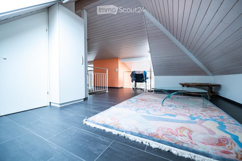 Modern attic room with slanted wooden ceilings, tiled floor, colorful rug, glass table, and chairs.