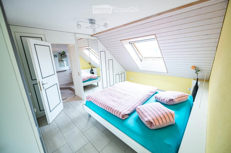 Bright attic bedroom with slanted wooden ceilings, large windows, and a bed with blue bedding and striped pillows. A mirror and wardrobe are visible, along with a vase of flowers on a surface.