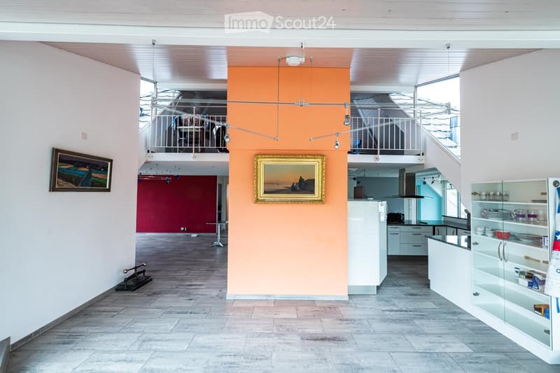 Open-plan living and kitchen area with modern white cabinets, stainless steel appliances, a kitchen island, and a staircase leading to an upper level. Features include a large framed picture on the wall, a small table and chair, and a staircase with a met