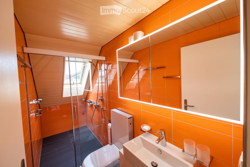 Modern bathroom with orange tiles, a large mirror with built-in lighting, a glass-enclosed shower, a white sink with a modern faucet, and a toilet.
