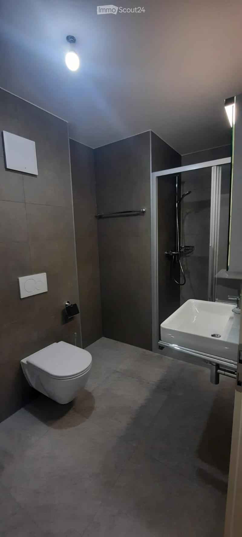 Modern bathroom with a toilet, sink, and walk-in shower with glass enclosure. Grey tiled walls and floor, recessed lighting, and a towel rack.