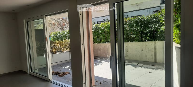 Open sliding glass doors lead to a tiled balcony area with a low concrete wall and surrounding greenery.