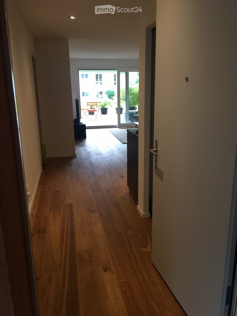 A well-lit hallway with wooden flooring, leading to a bright open area with large windows and a view of a balcony or terrace area.