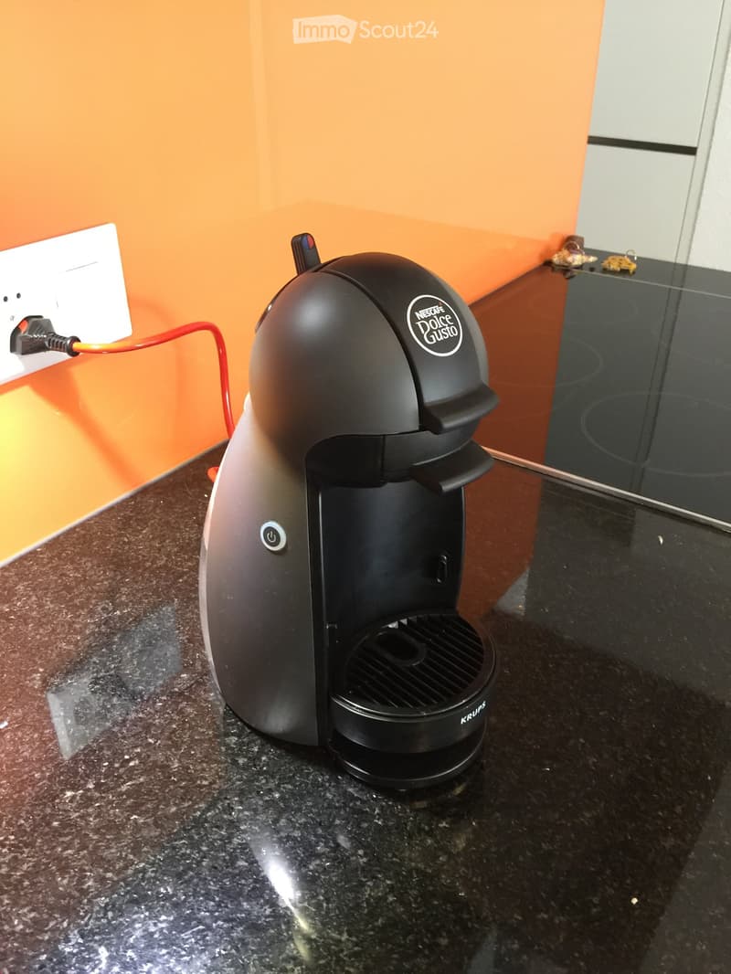 A black Nescafe Dolce Gusto coffee maker sits on a dark granite countertop, with an orange wall and a white electrical outlet in the background.