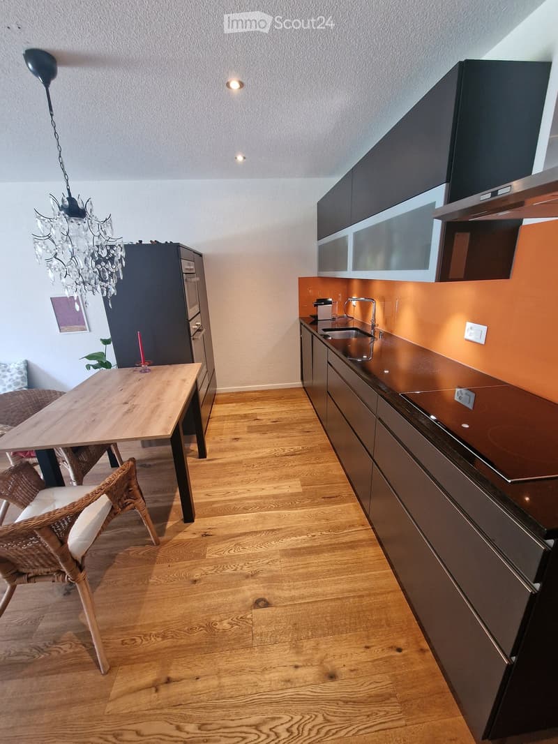 Modern kitchen with dark cabinets, wooden floor, and a small dining area with a table and chair. Features a chandelier, sink, and a candle on the table.