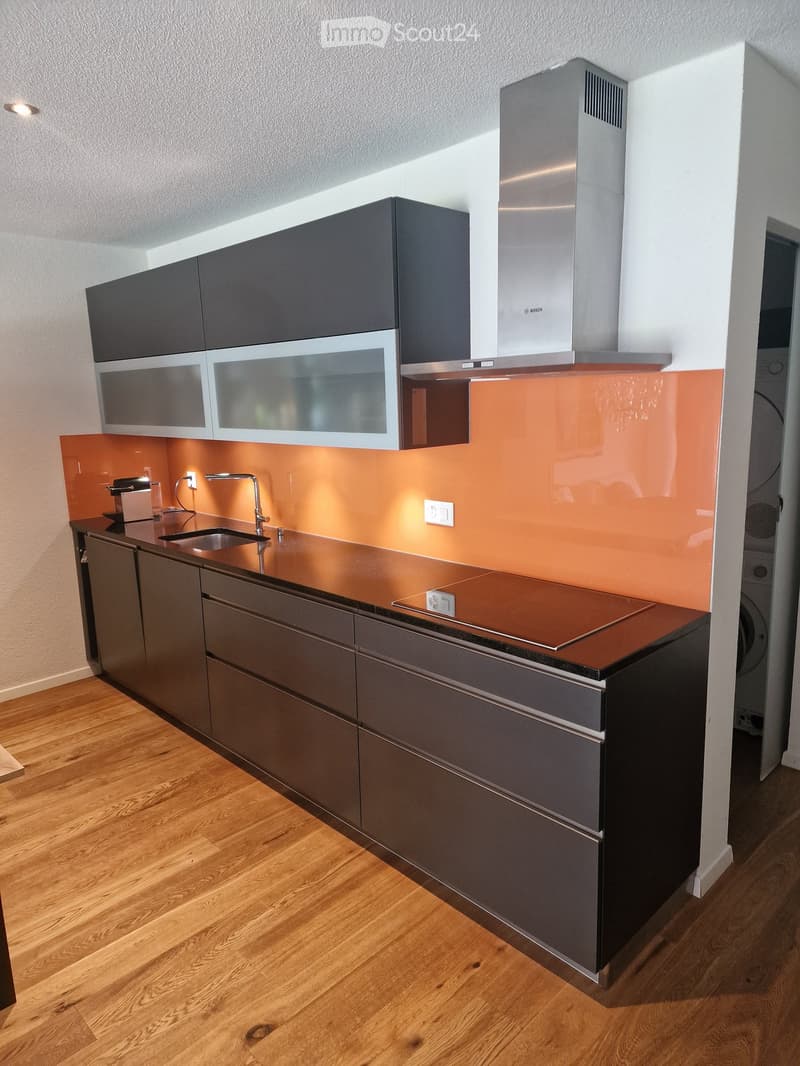 Modern kitchen with dark grey cabinets, stainless steel appliances, and a copper backsplash. Features include a sink, microwave, oven, and induction cooktop.
