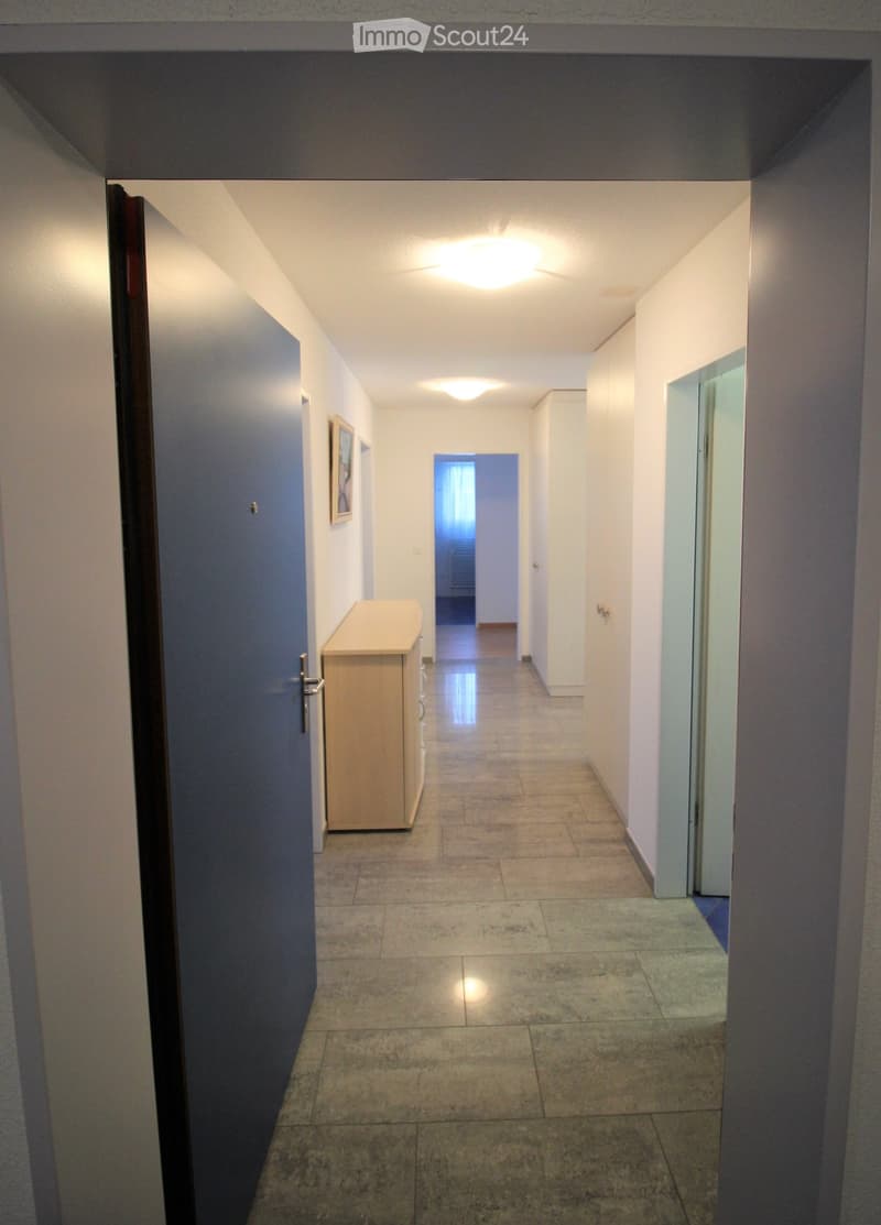 Empty hallway with tiled floor, white walls, and recessed lighting. A small cabinet is placed on the left side, and a framed picture hangs on the wall.