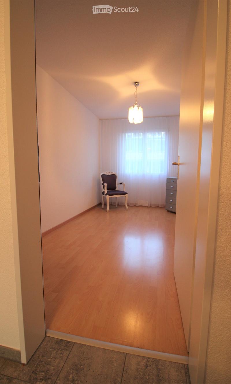 A small, well-lit room with wooden flooring, a single chair, and a cabinet. Natural light filters through a window with white curtains.