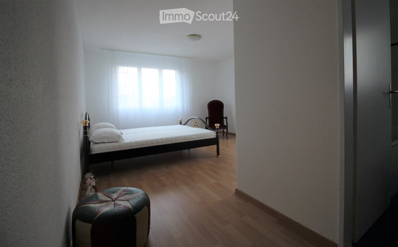 A small, well-lit bedroom with a wooden floor, a single bed with white bedding, a patterned leather stool, and a wooden chair near the window.