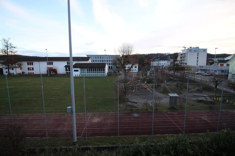 Outdoor area with a running track, playground equipment, grassy field, and surrounding buildings.