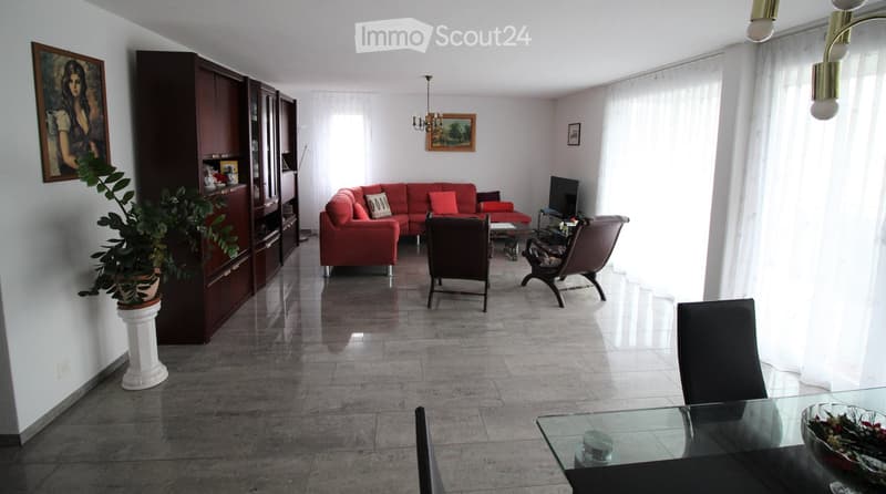 A spacious living room with a red sofa, wooden chairs, a glass dining table, a large cabinet, and a potted plant. The room features large windows with white curtains and a chandelier hanging from the ceiling.